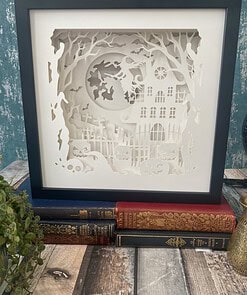 Framed Halloween-themed paper cut art with a haunted house, full moon, witch, and pumpkins, displayed on stacked books.