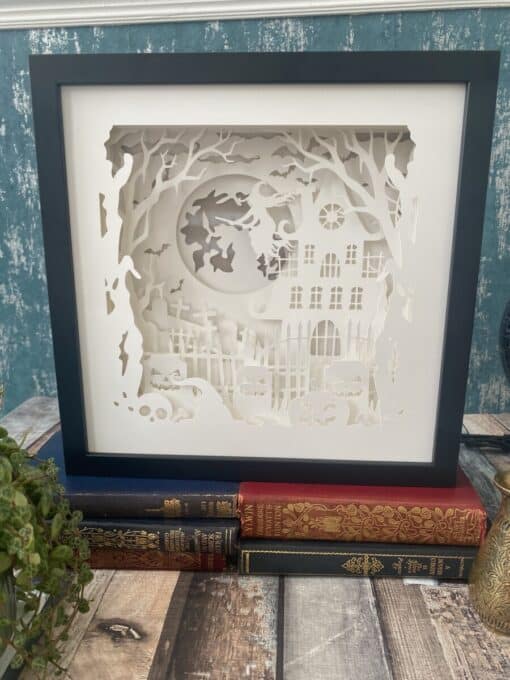 Framed Halloween-themed paper cut art with a haunted house, full moon, witch, and pumpkins, displayed on stacked books.