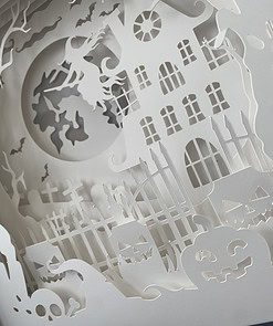 Intricate paper cut-out scene of a haunted house, graveyard, and jack-o'-lanterns with a witch flying on a broomstick under the moon.