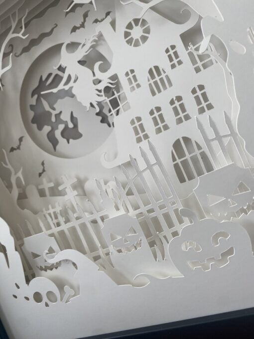 Intricate paper cut-out scene of a haunted house, graveyard, and jack-o'-lanterns with a witch flying on a broomstick under the moon.