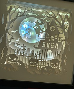 Shadow box art scene of a witch on broomstick, haunted house, full moon, bats, pumpkins, twisted trees, and cemetery gates.