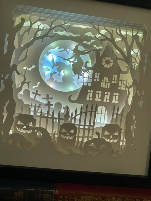 Shadow box art scene of a witch on broomstick, haunted house, full moon, bats, pumpkins, twisted trees, and cemetery gates.