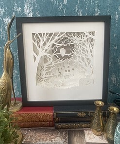 Framed paper cut art of a spooky graveyard scene with an owl, displayed on ornate books, beside brass bird and vases.