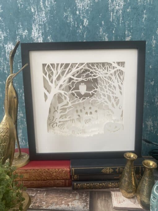 Framed paper cut art of a spooky graveyard scene with an owl, displayed on ornate books, beside brass bird and vases.