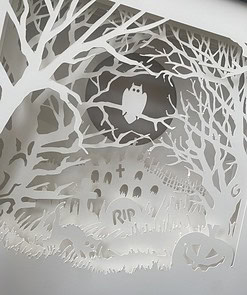 A paper cutout scene featuring an owl in a tree, gravestones, a black cat, and a jack-o'-lantern, representing a spooky graveyard.