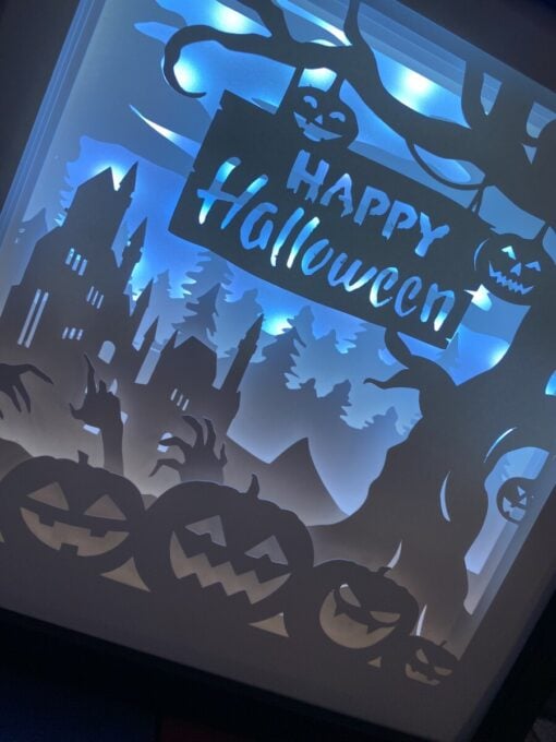 Illuminated Halloween scene with pumpkins, a haunted house, spooky hands, trees, and a "Happy Halloween" sign against a blue background.