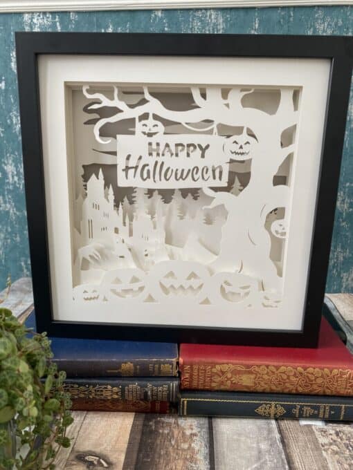 Framed Halloween paper art with "Happy Halloween" sign, pumpkins, haunted house, and tree, set on a stack of vintage books.