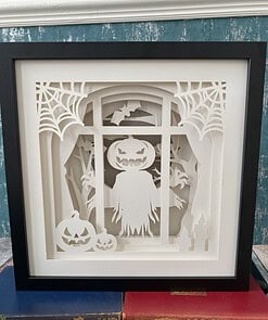 Paper art of a ghost with a pumpkin head, surrounded by spider webs, bats, pumpkins, and candles in a black frame.