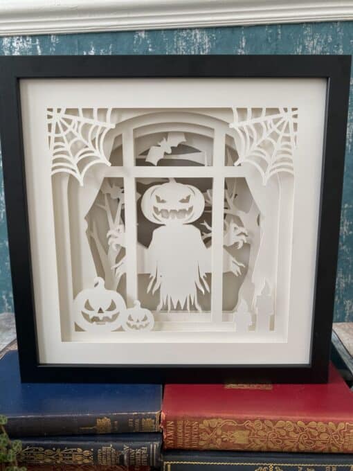 Paper art of a ghost with a pumpkin head, surrounded by spider webs, bats, pumpkins, and candles in a black frame.