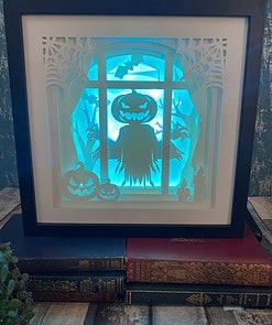 Halloween-themed shadowbox with glowing pumpkins, a scarecrow, bats, and trees in a window frame, placed on top of vintage books.