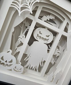 Paper cutout depicting a spooky Halloween scene with a pumpkin-headed scarecrow, bats, and carved pumpkins in front of a window.