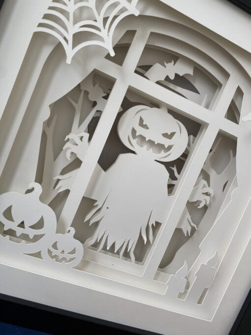 Paper cutout depicting a spooky Halloween scene with a pumpkin-headed scarecrow, bats, and carved pumpkins in front of a window.