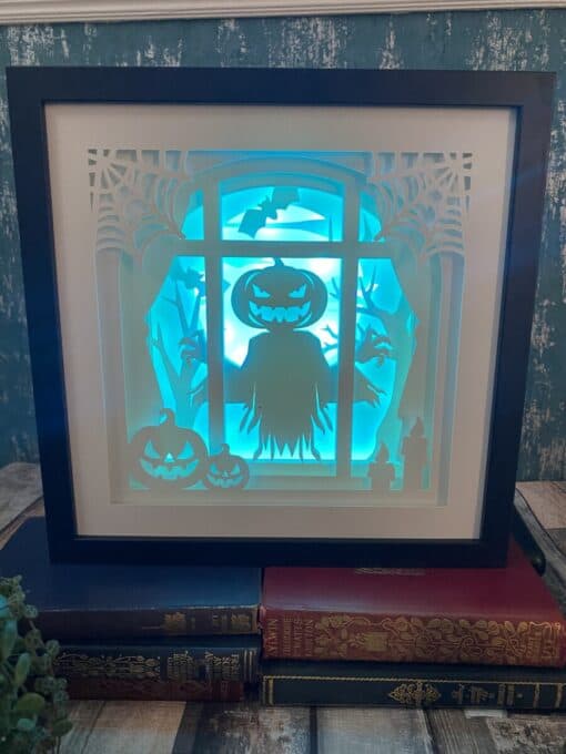 Halloween-themed shadowbox with glowing pumpkins, a scarecrow, bats, and trees in a window frame, placed on top of vintage books.