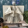 DIY paper angel decoration made from book pages, set on a wooden table with pine cones and plants, colorful lights in the background.