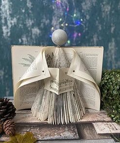 DIY paper angel decoration made from book pages, set on a wooden table with pine cones and plants, colorful lights in the background.