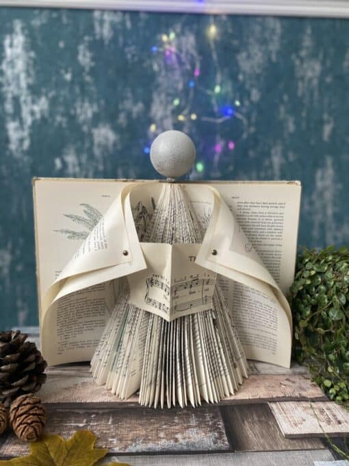 DIY paper angel decoration made from book pages, set on a wooden table with pine cones and plants, colorful lights in the background.