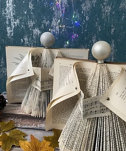 Folded book pages crafted into decorative trees with glittery baubles on top, set against a teal wall with fairy lights.