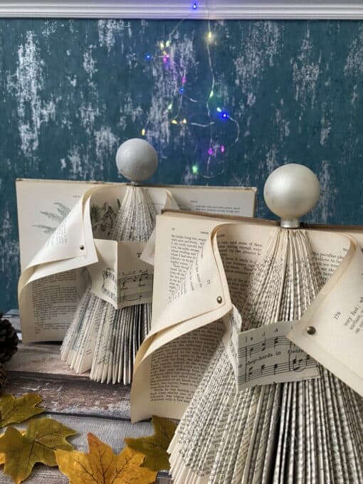 Folded book pages crafted into decorative trees with glittery baubles on top, set against a teal wall with fairy lights.