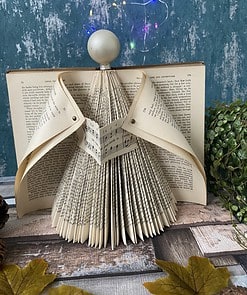 Book pages folded into an angel shape with a spherical head, set against a textured blue wall and colorful string lights.