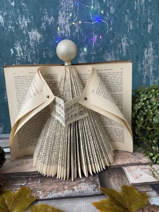 Book pages folded into an angel shape with a spherical head, set against a textured blue wall and colorful string lights.