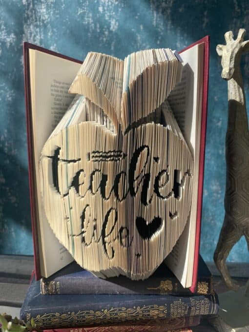 Book pages folded to form the shape of an apple with the words "teacher life" and a heart carved into them, placed on two other books.