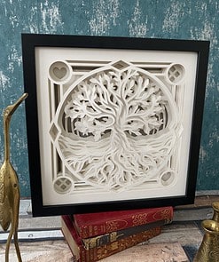 Framed white paper-cut art of a tree with roots, displayed on vintage red books, against a teal textured wall.