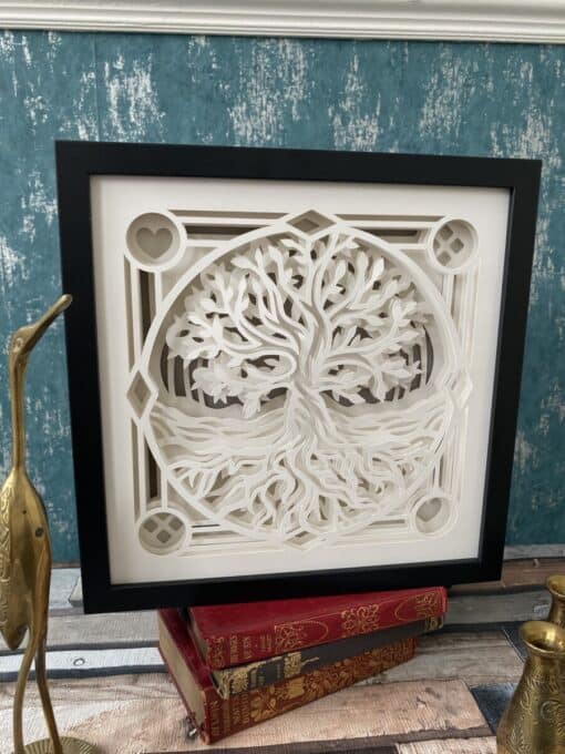 Framed white paper-cut art of a tree with roots, displayed on vintage red books, against a teal textured wall.