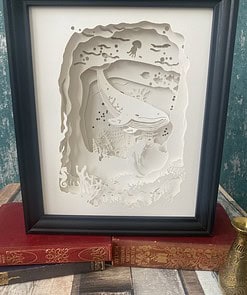 Framed paper art depicting an underwater scene with a whale and sea creatures, displayed on vintage books.