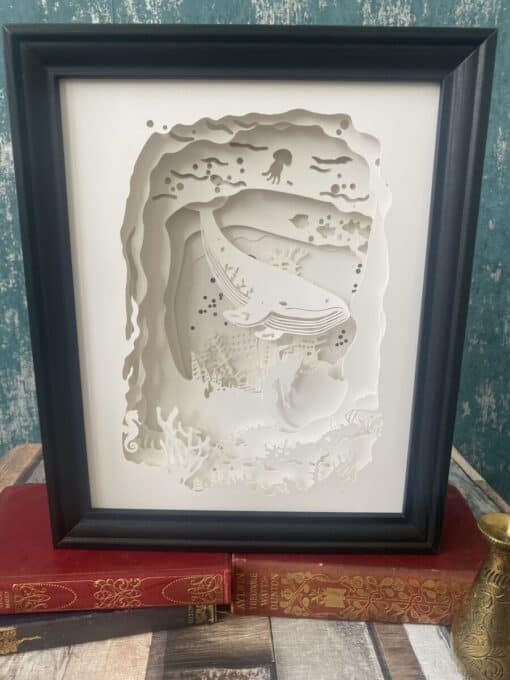 Framed paper art depicting an underwater scene with a whale and sea creatures, displayed on vintage books.