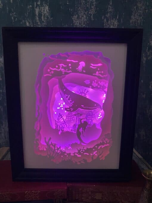 Framed luminous sea scene with a whale and coral, glowing in purple and pink hues against a dark background.