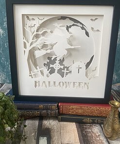 Framed Halloween art with a witch on a broomstick, bats, and a graveyard scene, sitting on vintage books.