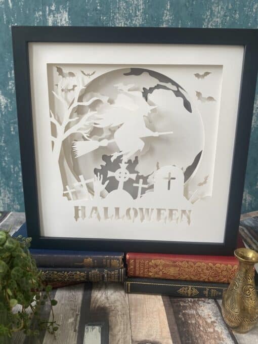 Framed Halloween art with a witch on a broomstick, bats, and a graveyard scene, sitting on vintage books.