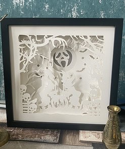 Framed paper art with a Halloween theme depicting witches, cauldron, skulls, bats, and spooky trees in intricate white cutouts.