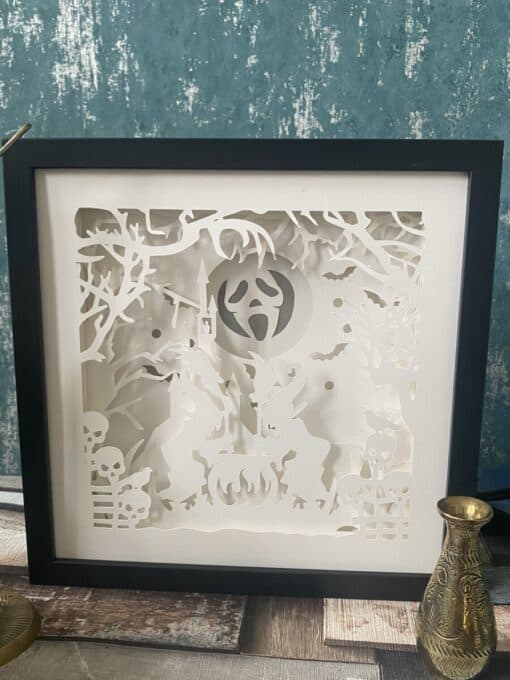 Framed paper art with a Halloween theme depicting witches, cauldron, skulls, bats, and spooky trees in intricate white cutouts.