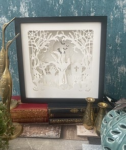 Framed intricate paper cut art of a Halloween scene with trees, bats, and tombstones, surrounded by vintage decor and books.