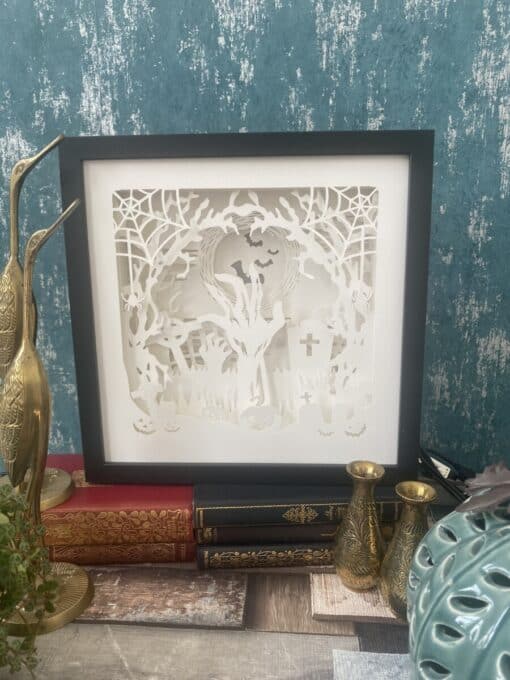 Framed intricate paper cut art of a Halloween scene with trees, bats, and tombstones, surrounded by vintage decor and books.