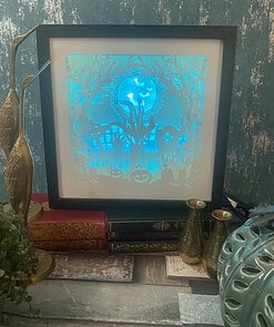 Framed Halloween-themed backlit artwork on a shelf, surrounded by vintage books and gold decorative items.