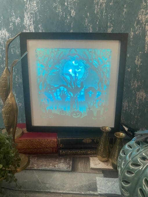 Framed Halloween-themed backlit artwork on a shelf, surrounded by vintage books and gold decorative items.