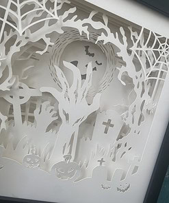 Intricate Halloween-themed paper cut-out art featuring pumpkins, tombstones, tree branches, and bats against a full moon.