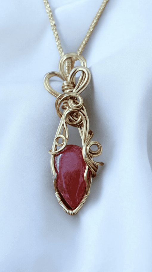 Gold pendant with intricate wire design enclosing a reddish gemstone, hanging from a twisted gold chain, on a white background.