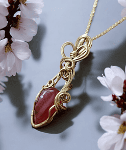 Gold wire-wrapped red gemstone pendant with intricate swirls, surrounded by soft white flowers on a light gray background.