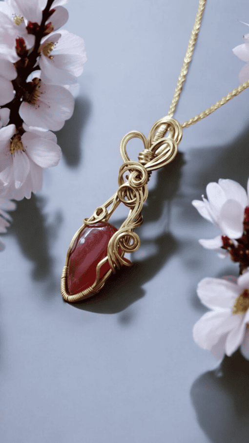 Gold wire-wrapped red gemstone pendant with intricate swirls, surrounded by soft white flowers on a light gray background.