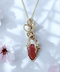 Gold wire-wrapped pendant with a red gemstone on a gold chain, surrounded by soft pink flowers against a white background.