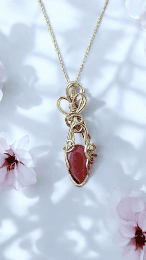 Gold wire-wrapped pendant with a red gemstone on a gold chain, surrounded by soft pink flowers against a white background.