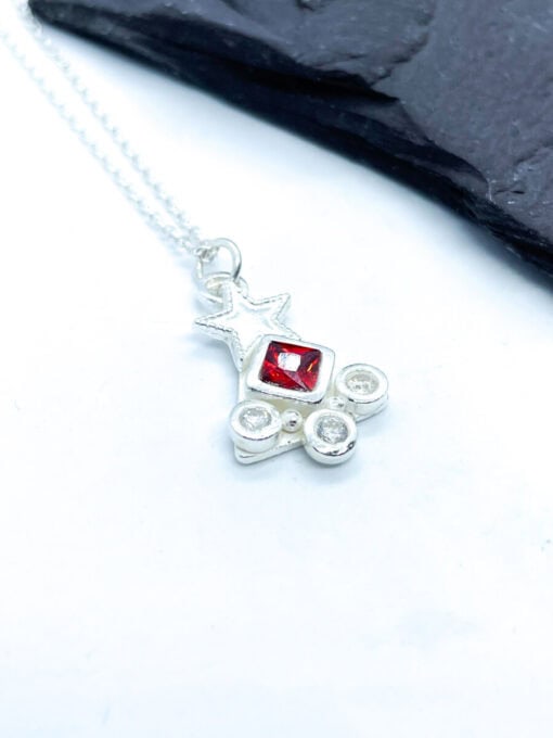 Silver necklace pendant with a star shape, featuring a central red gem and two smaller clear crystals, displayed on a white background.