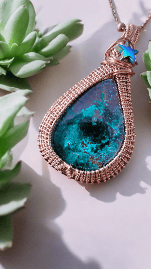 Close-up of a handcrafted teardrop pendant with a blue-green stone set in intricate wire wrapping, surrounded by succulents.