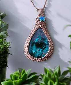 Teardrop-shaped pendant with a turquoise stone, wrapped in copper wire, and a blue star charm, surrounded by green plants.