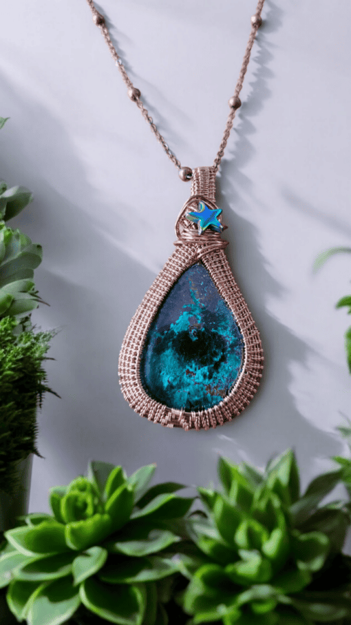 Teardrop-shaped pendant with a turquoise stone, wrapped in copper wire, and a blue star charm, surrounded by green plants.