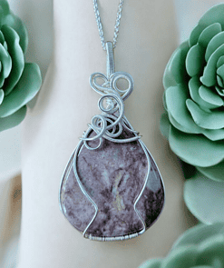 Silver wire-wrapped pendant with a purple stone on a chain, surrounded by green succulents in the background.