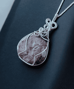 Close-up of a pendant necklace with an intricate silver wire design encasing a teardrop-shaped purple stone, on a dark fabric background.